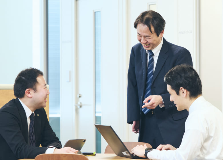 会議室で資料を囲んで打ち合わせをする右上肢スポーツベットio つながらないの男性社員と同僚たち。身振り手振りを交えてフランクに会話する業務風景の写真